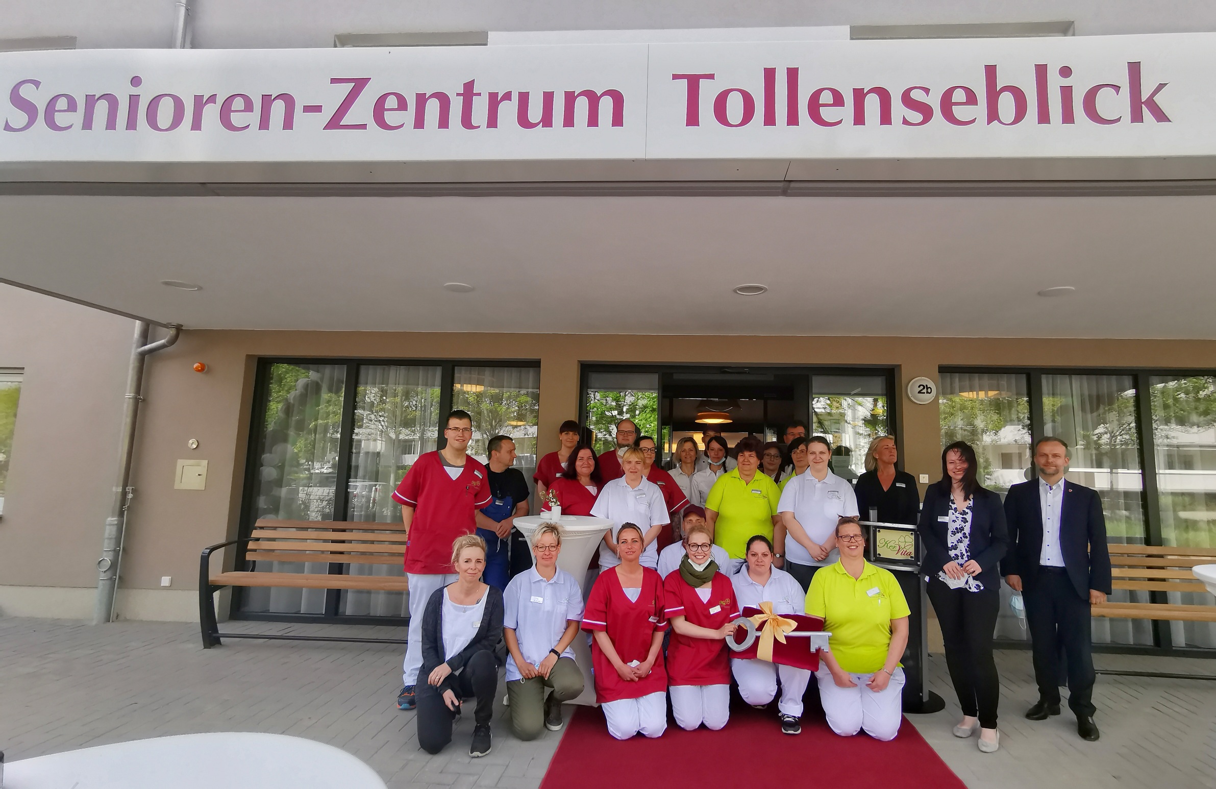 Gruppenfoto vor dem Eingang des „Senioren-Zentrums Tollenseblick“. Pflegekräfte in farbiger Dienstkleidung sowie weitere Mitarbeitende und Gäste stehen und knien lächelnd auf einem roten Teppich vor dem Gebäude – die Szene wirkt festlich und gemeinschaftl